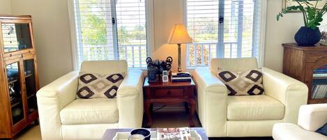 A living room with two white leather sofas, a wooden coffee table with magazines and a glass, and a lamp on a side table.