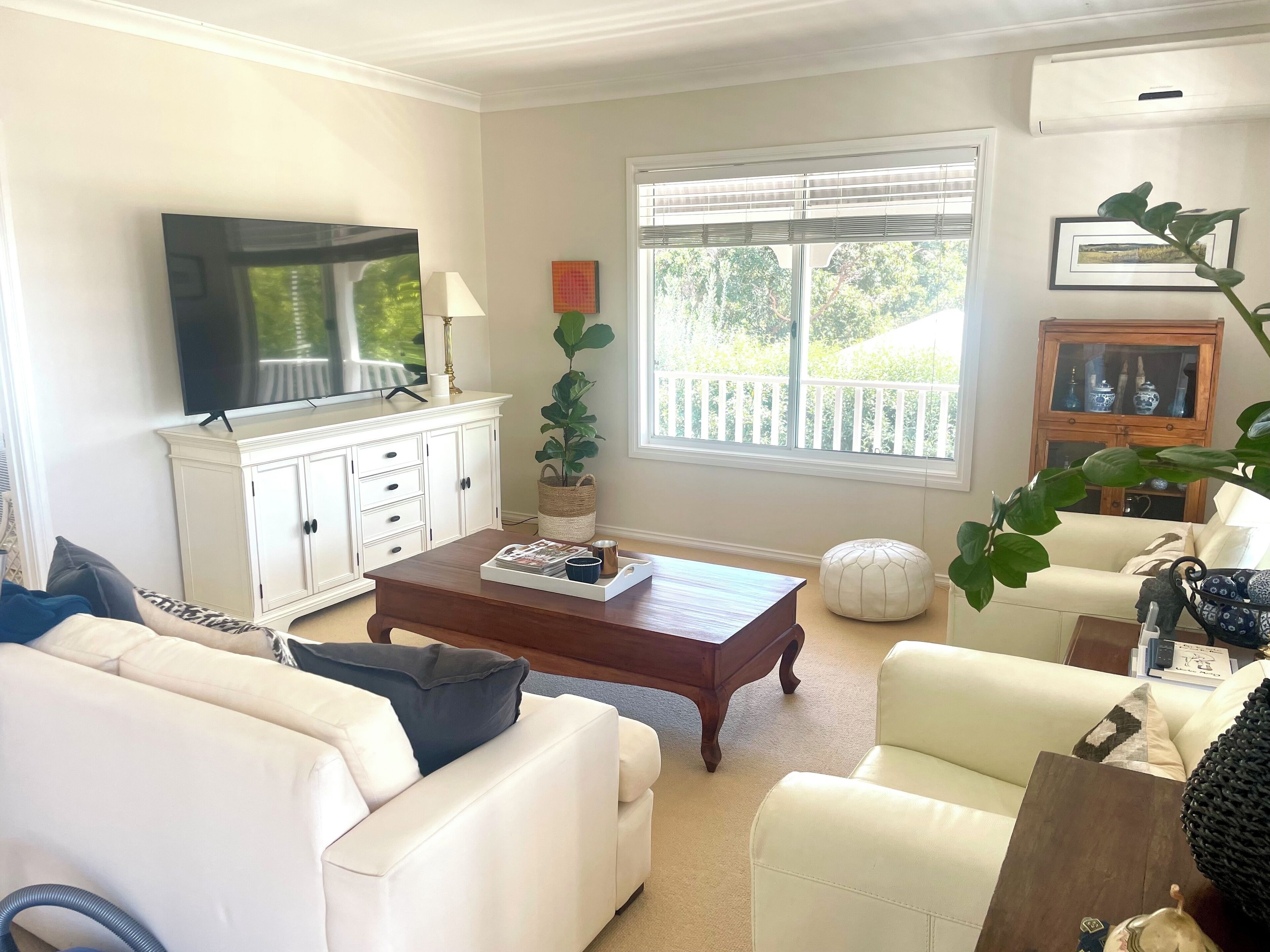 A living room with a TV, white furniture, a coffee table, and a large window with a view of greenery.