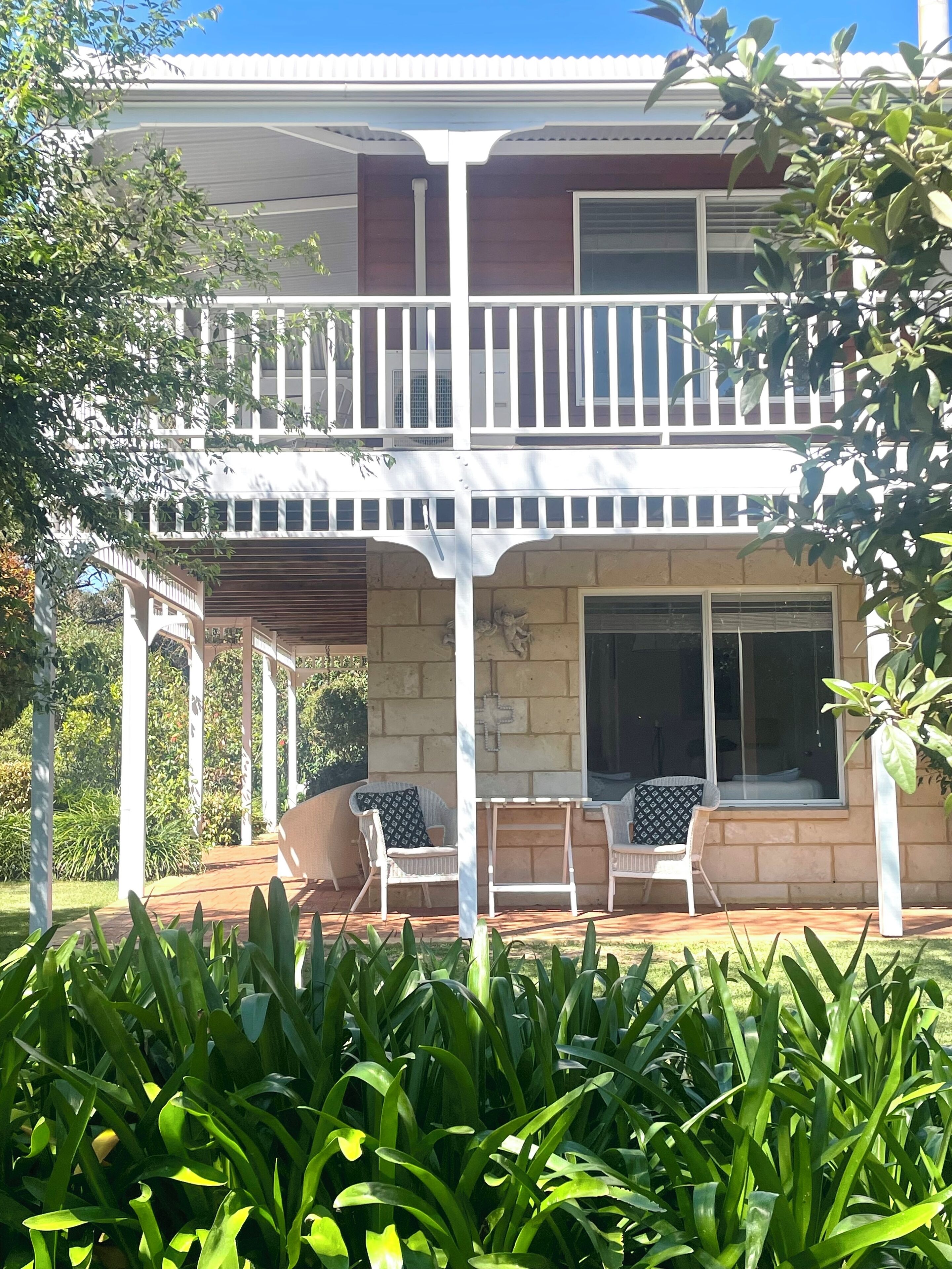 A two-story house with a balcony, a covered patio with chairs, and a garden in the foreground.