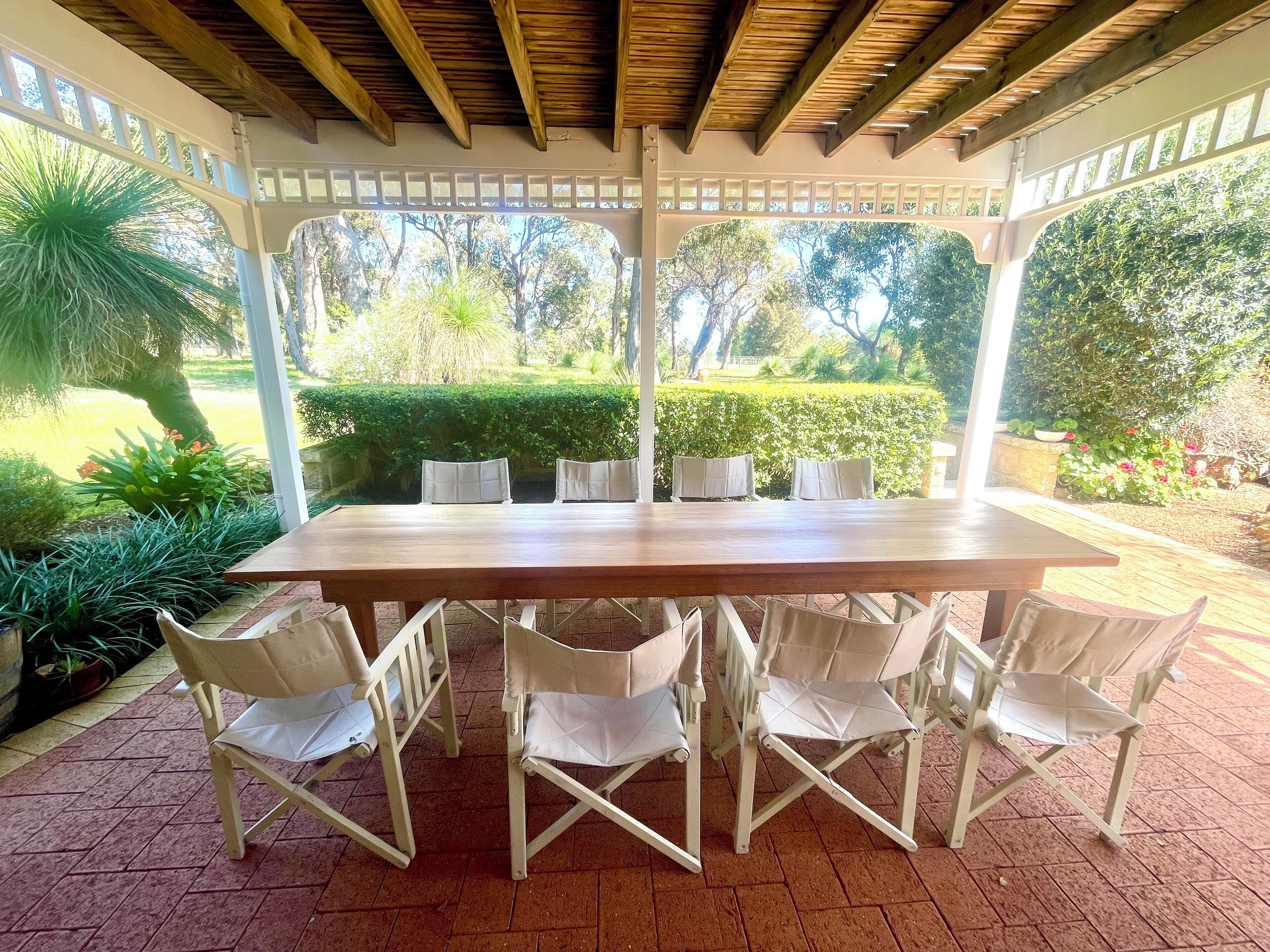 A covered outdoor dining area with a wooden table and white chairs, surrounded by greenery and a view of the garden.