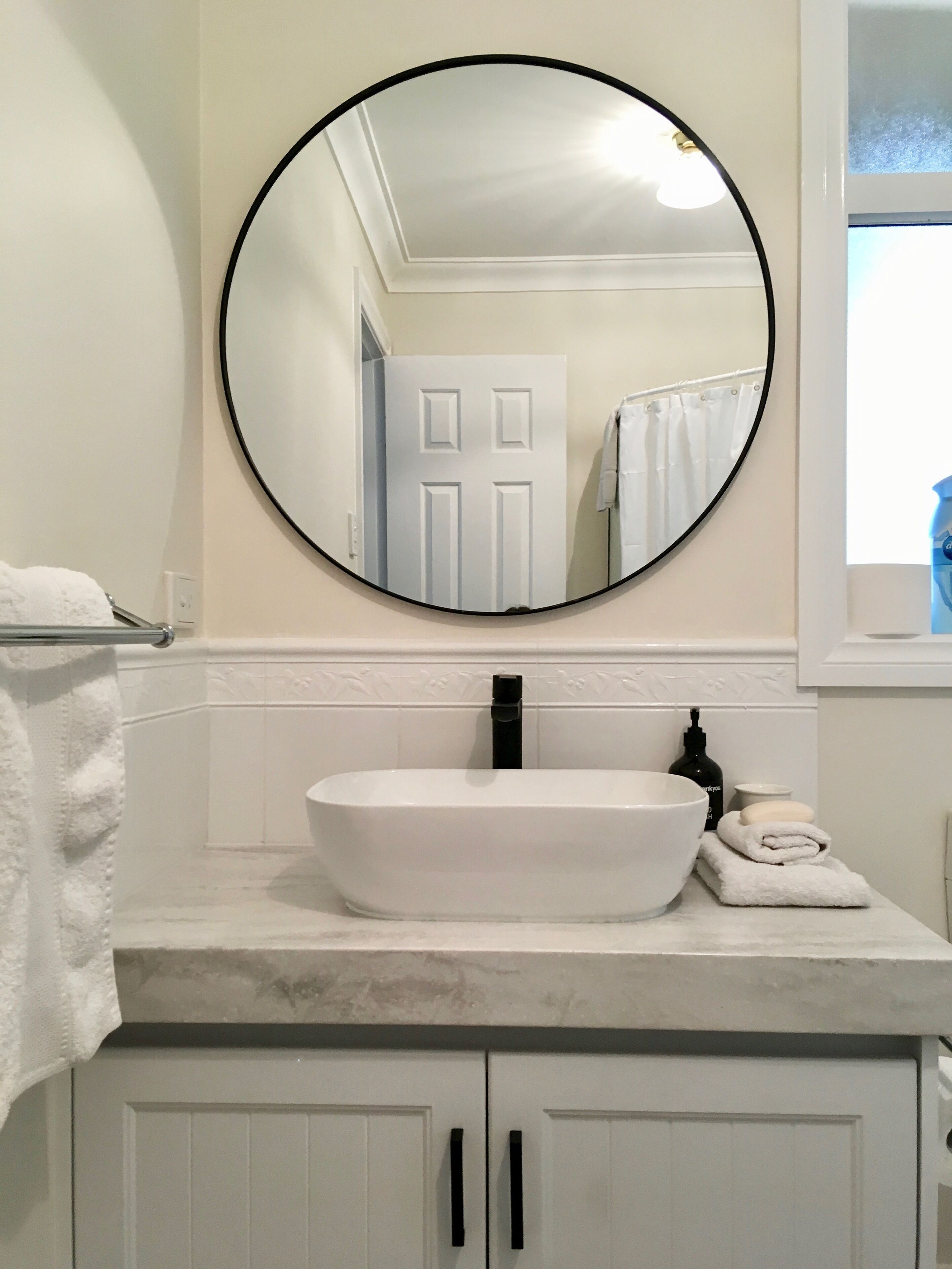 A bathroom with a round mirror, a white sink, and a window with a view of the outside.