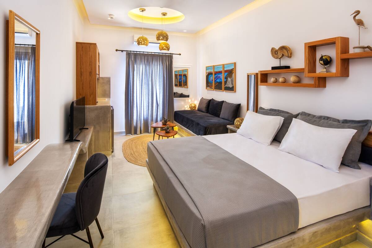 A modern hotel room with a large bed, a desk, and a chair. There is a TV, a small table, and a shelf with decorative items.