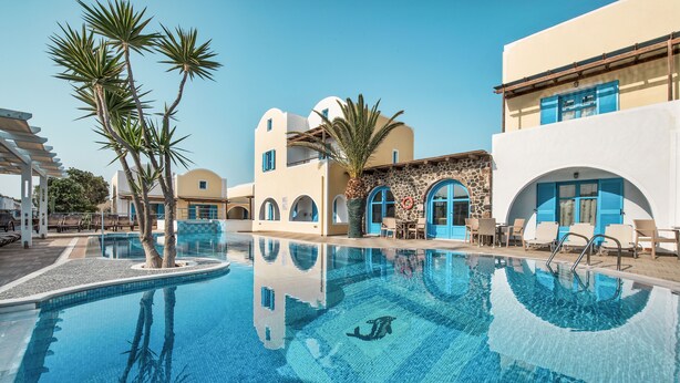 Zona de piscina amb aigua cristal·lina blava, palmeres i un edifici amb portes i finestres blaves.