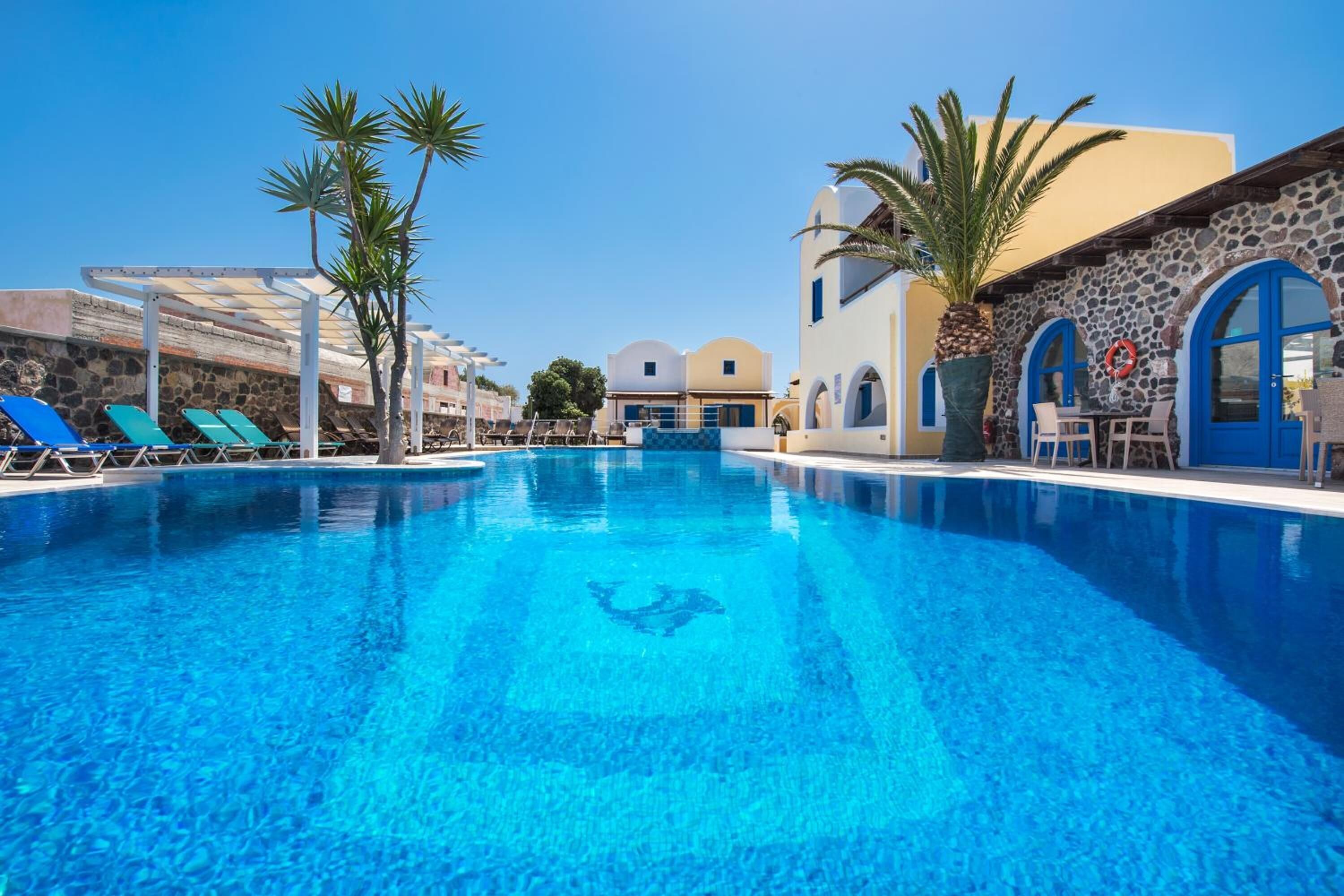 Piscina azul cristalina con sillas de descanso y palmeras, rodeada de edificios de piedra con puertas azules.