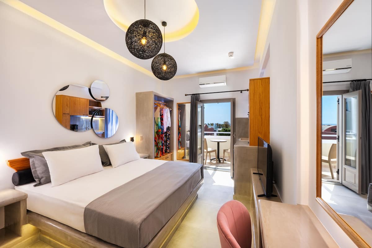 A modern hotel room with a large bed, a desk with a chair, a TV, a wardrobe, and a balcony with a view.