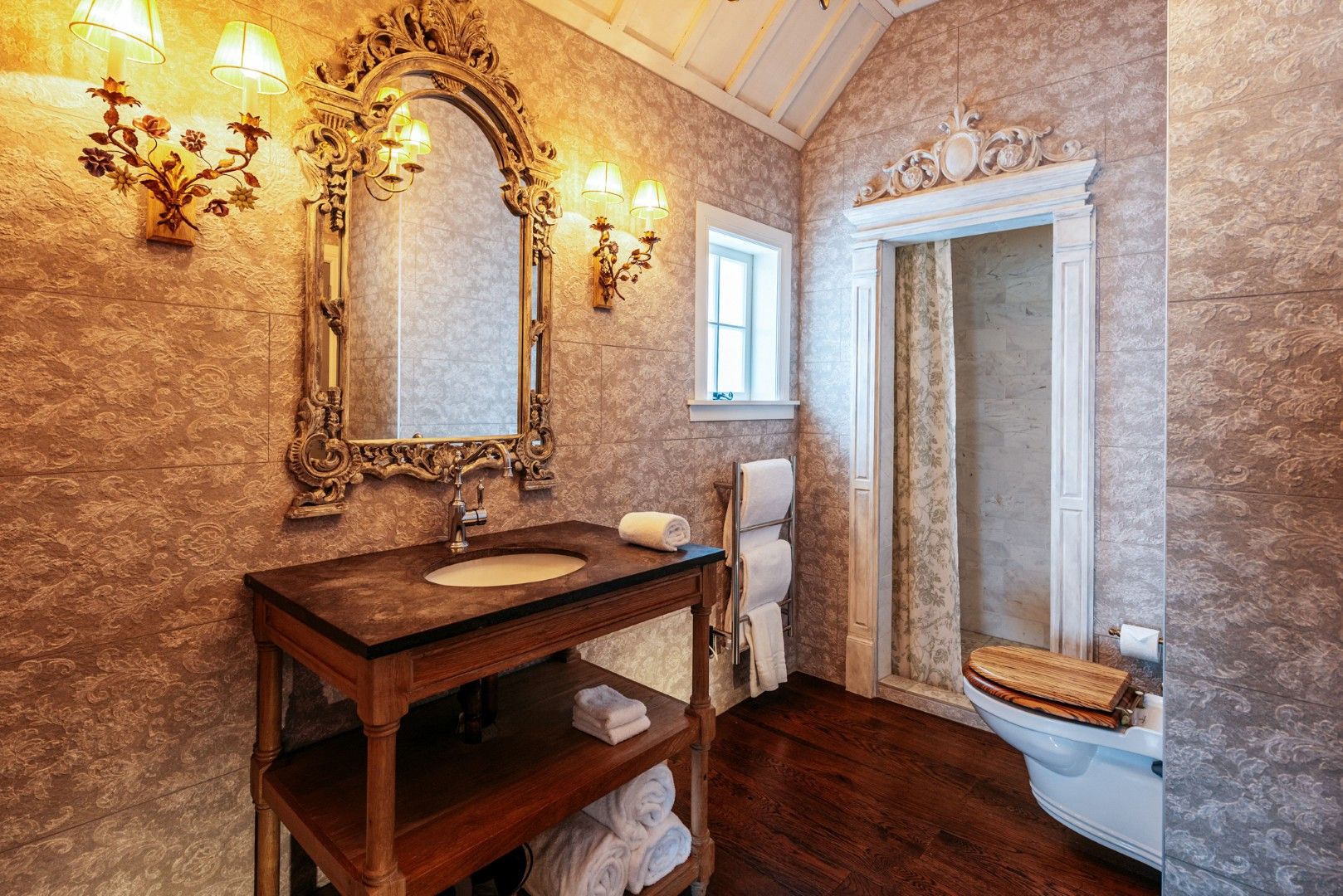 A bathroom with a wooden vanity, a large ornate mirror, wall sconces, a towel rack, and a toilet.