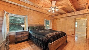 Ein Schlafzimmer einer Holzhütte mit Bett, Nachttischen, Deckenventilator und einem Fenster mit Jalousien.