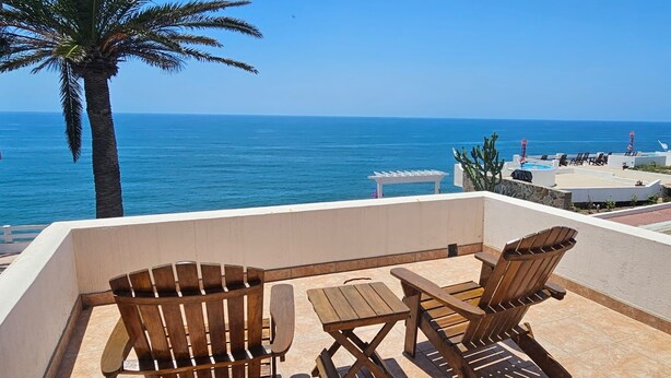 Eine Terrasse mit Holzmöbeln, einer Palme und Blick aufs Meer.