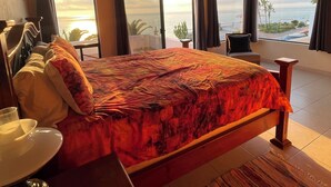 Ein Schlafzimmer mit einem großen Bett, einem Sessel und Meerblick durch große Fenster.
