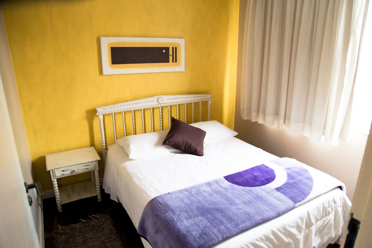 Un dormitori amb un llit, una tauleta de nit, un calefactor fixat a la paret i una finestra amb cortines.