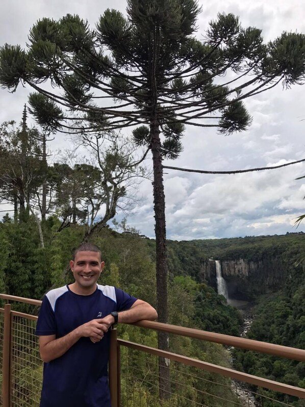 A man standing on a balcony with a railing, overlooking a lush forest and a waterfall.