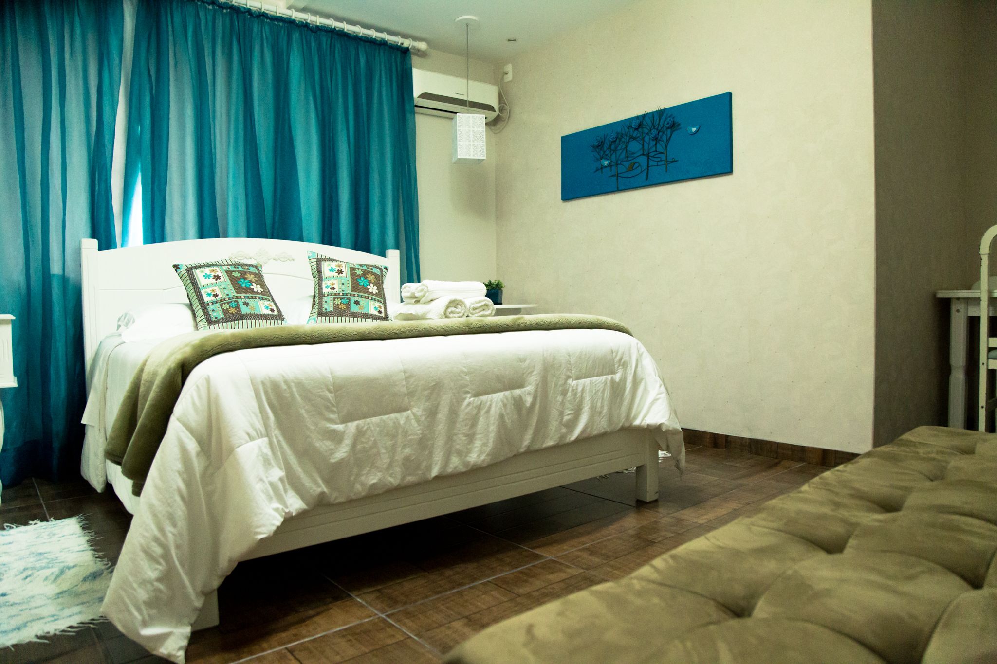 A bedroom with a bed, a bedside table, a window with curtains, a wall-mounted artwork, and a wooden floor.