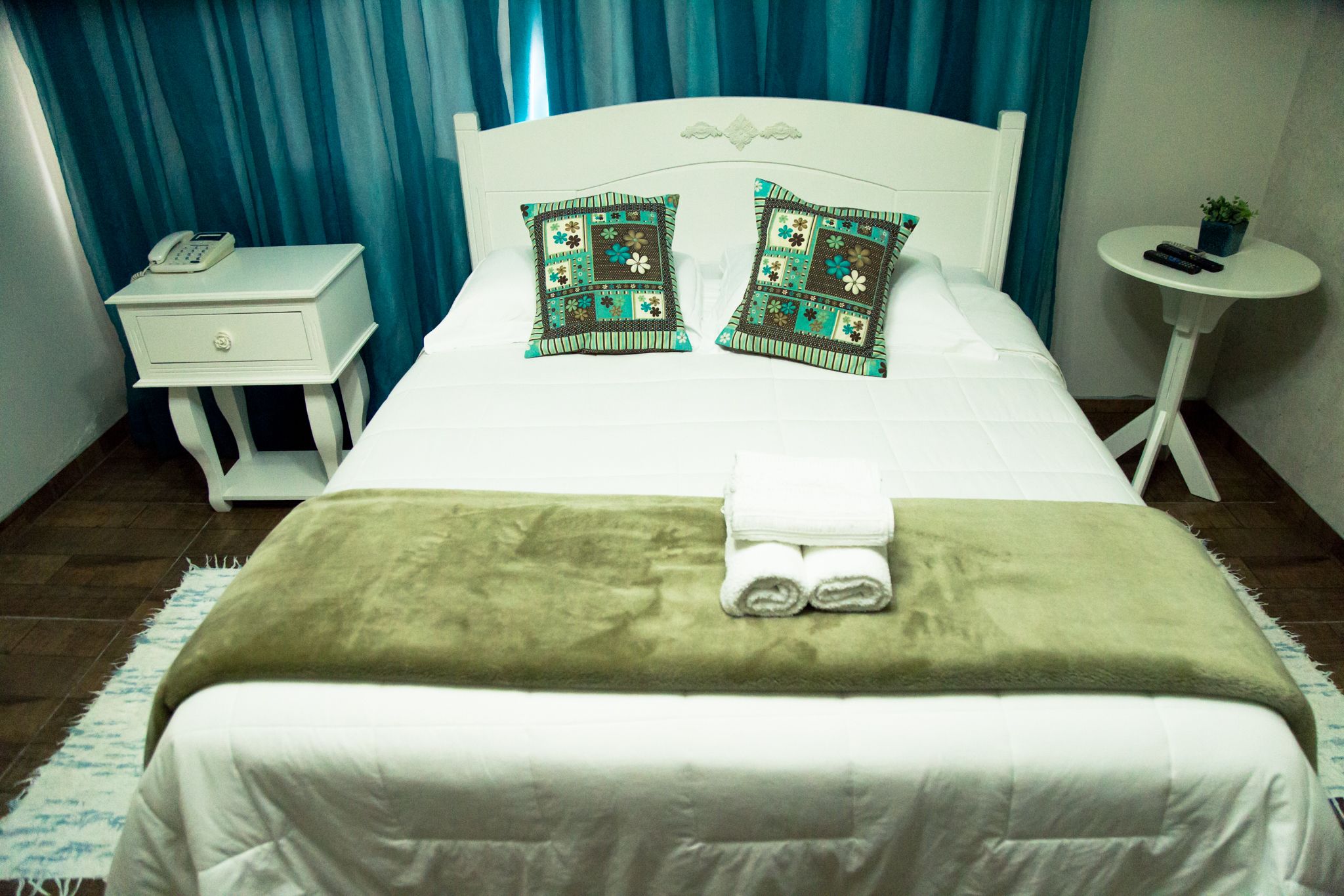 Una cama bien hecha con una manta verde y dos almohadas, una mesita de noche con un teléfono, una pequeña mesa con una planta y una cortina azul al fondo.