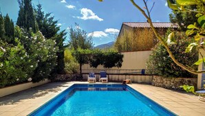 A rectangular swimming pool with a tiled deck, surrounded by lush greenery and a stone wall.