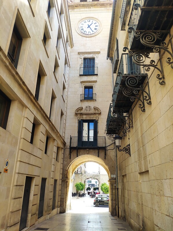 A narrow alleyway with a clock tower, stone buildings, and an archway leading to a plaza.