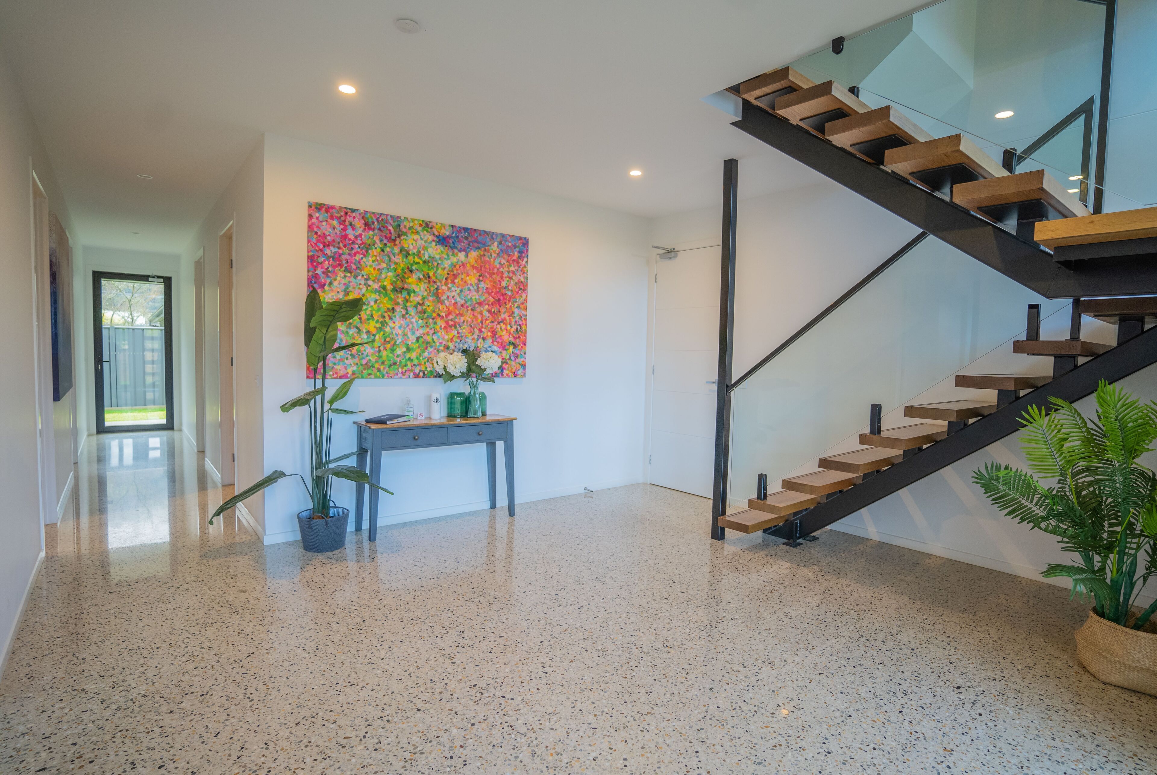 Un intérieur moderne avec un escalier, un tableau abstrait aux couleurs vives, un bureau agrémenté d’une plante, et une plante en pot près de l’escalier.