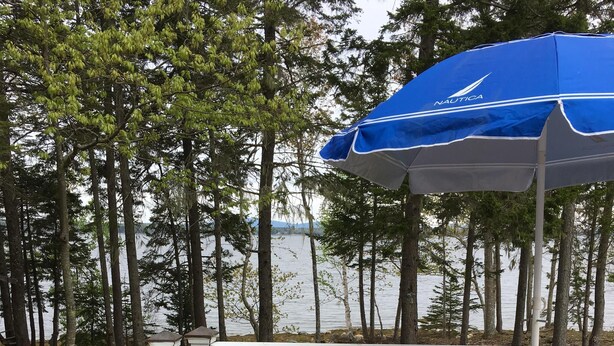 Une terrasse en bois avec un fauteuil Adirondack blanc et un parasol bleu, donnant sur un lac bordé d’arbres.