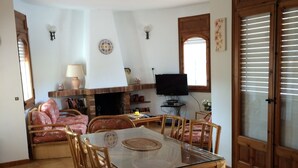 Ein Essbereich mit Glastisch und Korbstühlen, einem Kamin, einem Fernseher und einem großen Fenster mit Jalousien.