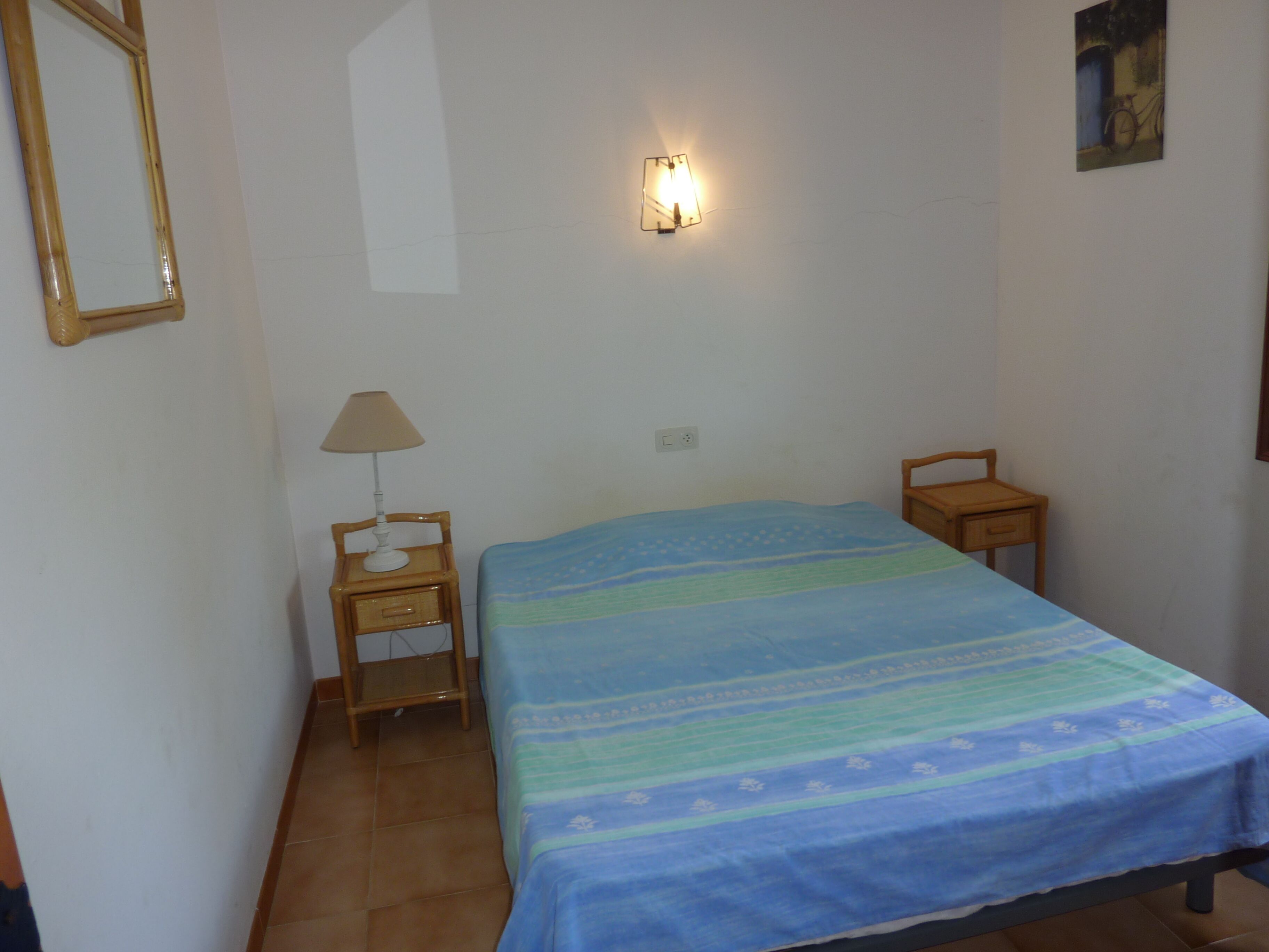 A bedroom with a bed, a nightstand with a lamp, a wall-mounted light, and a small wooden chair.