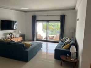 A living room with a large TV, two sofas, a coffee table, and a sliding glass door leading to an outdoor seating area.