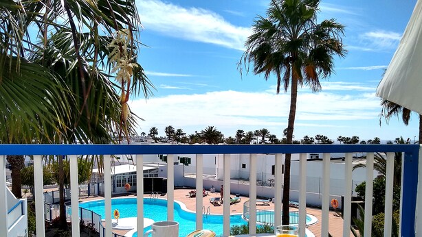 A balcony with a view of a swimming pool, a table with breakfast items, and a white railing. - cosy appartment in a residencial development near the sea (Puerto del Carmen)