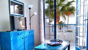 A room with a blue cabinet, a TV, a table with a decorative plate, and a view of palm trees through large windows. - cosy appartment in a residencial development near the sea (Puerto del Carmen)
