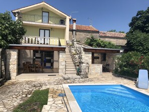 Una casa de dos pisos con una escalera de piedra que conduce a un balcón, una piscina y un patio amueblado al aire libre.