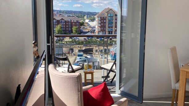 A modern room with a large glass door leading to an outdoor area. Inside, there is a beige armchair with a red pillow, a small wooden table with a drink, and a chair. The room has a tiled floor and a view of buildings and a parking lot outside.