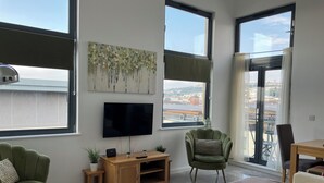 A modern living room with a flat-screen TV, green armchairs, a wooden TV stand, and large windows offering a city view.