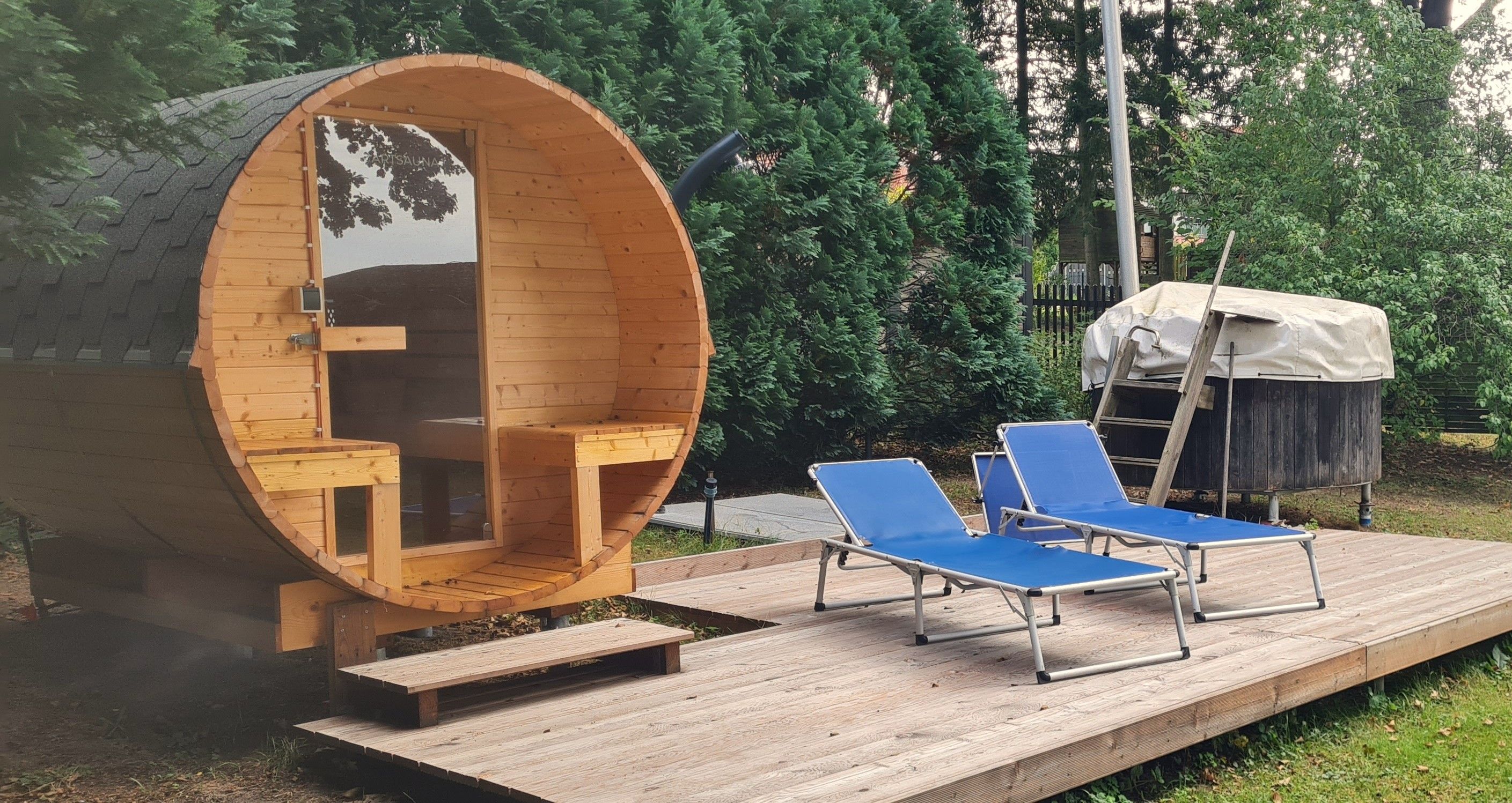 Eine baumstamähnliche Holzkonstruktion mit einer Bank und einem kleinen Tisch, zwei blaue Lounge-Stühle und einem überdachten Bereich mit einer Leiter.