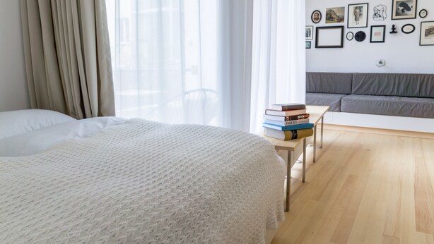Ein modernes Schlafzimmer mit einem großen Bett, einem Nachttisch mit Büchern, einem grauen Sofa und einer Wand mit gerahmten Bildern.