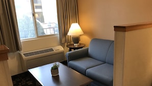 A hotel room with a blue sofa, a small table with a plant, a lamp, and a window with curtains.