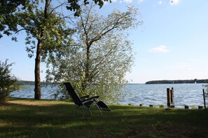 Un paisaje junto al lago con una silla bajo un árbol, un muelle de madera y un barco distante en el agua.