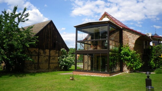 A modern building with a glass facade, a brick structure, and a wooden barn in the background. - Studio with winter garden and lake view - Ferienhof Löschenrand (Bad Saarow)