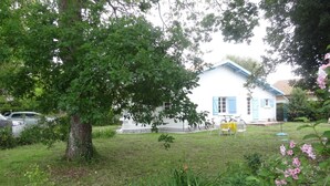 Un bâtiment blanc avec des volets bleus, un jardin clôturé et un arbre au premier plan.