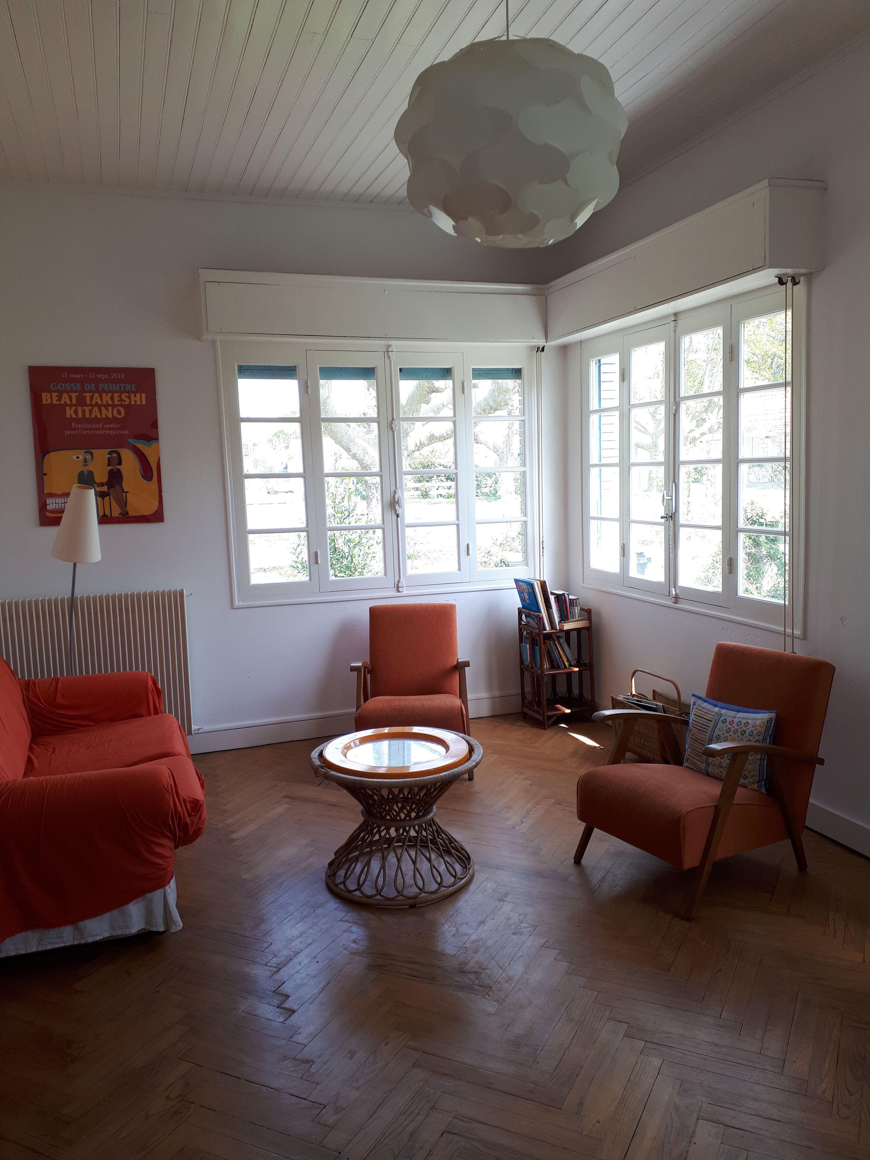 Une pièce dotée d’un parquet, de deux fauteuils rouges, d’une table ronde et de grandes fenêtres.