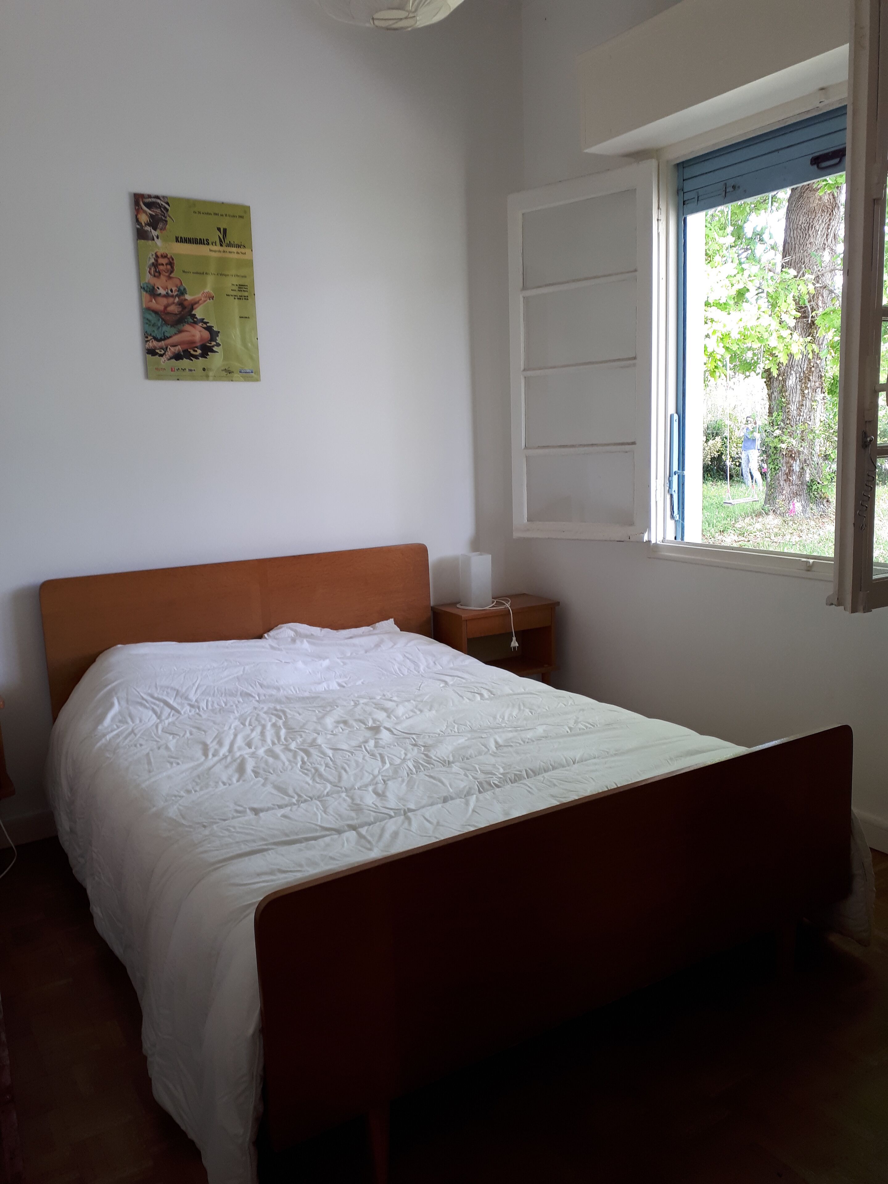 A bedroom with a wooden bed, a nightstand, a window with a view of greenery, and a poster on the wall.