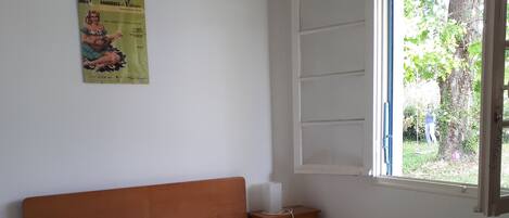 A bedroom with a wooden bed, a nightstand, a window with a view of greenery, and a poster on the wall.