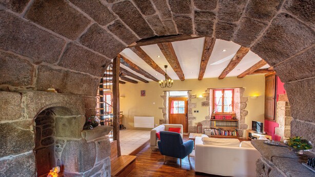 Una habitación de paredes de piedra con chimenea, vigas de madera, un sofá blanco y un sillón azul.