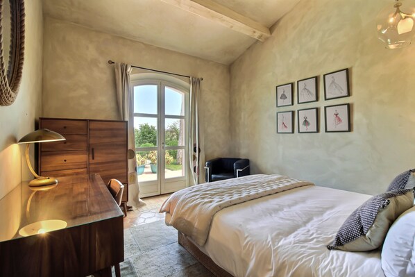 A bedroom with a large bed, a wooden desk, a chair, a window with curtains, and framed artwork on the wall.