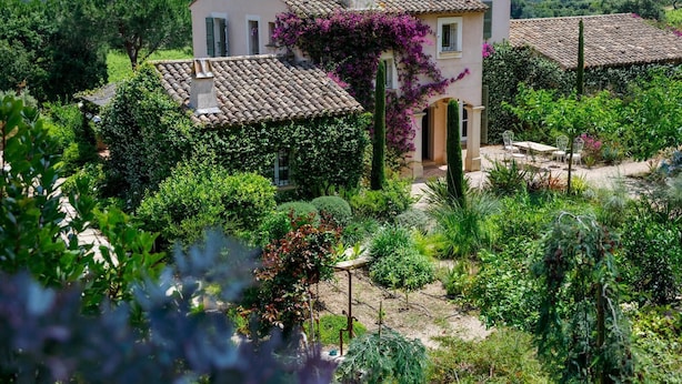 Una casa de dos pisos con techo de tejas, rodeada de abundante vegetación y plantas en flor.