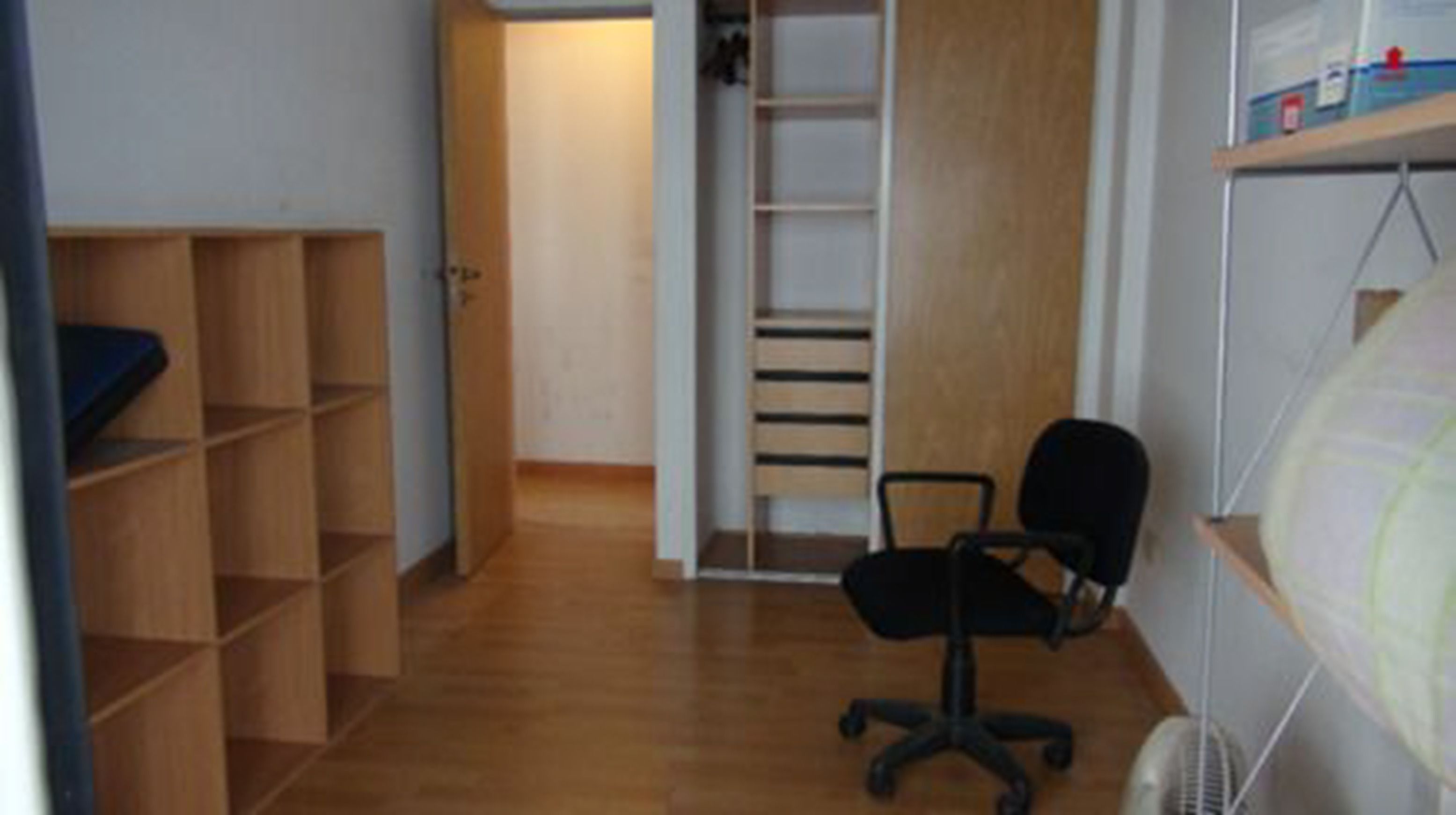 A compact room with a wooden floor, a black office chair, a white shelving unit, and a door leading to another room.