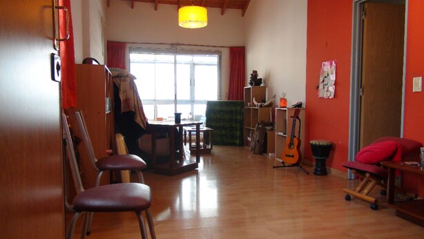 A room with wooden floors, a large window with red curtains, a guitar, and a chair. - Sunny 2 Bedroom Apartment (Buenos Aires)