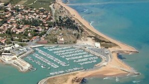 An aerial view of a coastal marina with boats docked, a sandy beach, and a residential area nearby.
