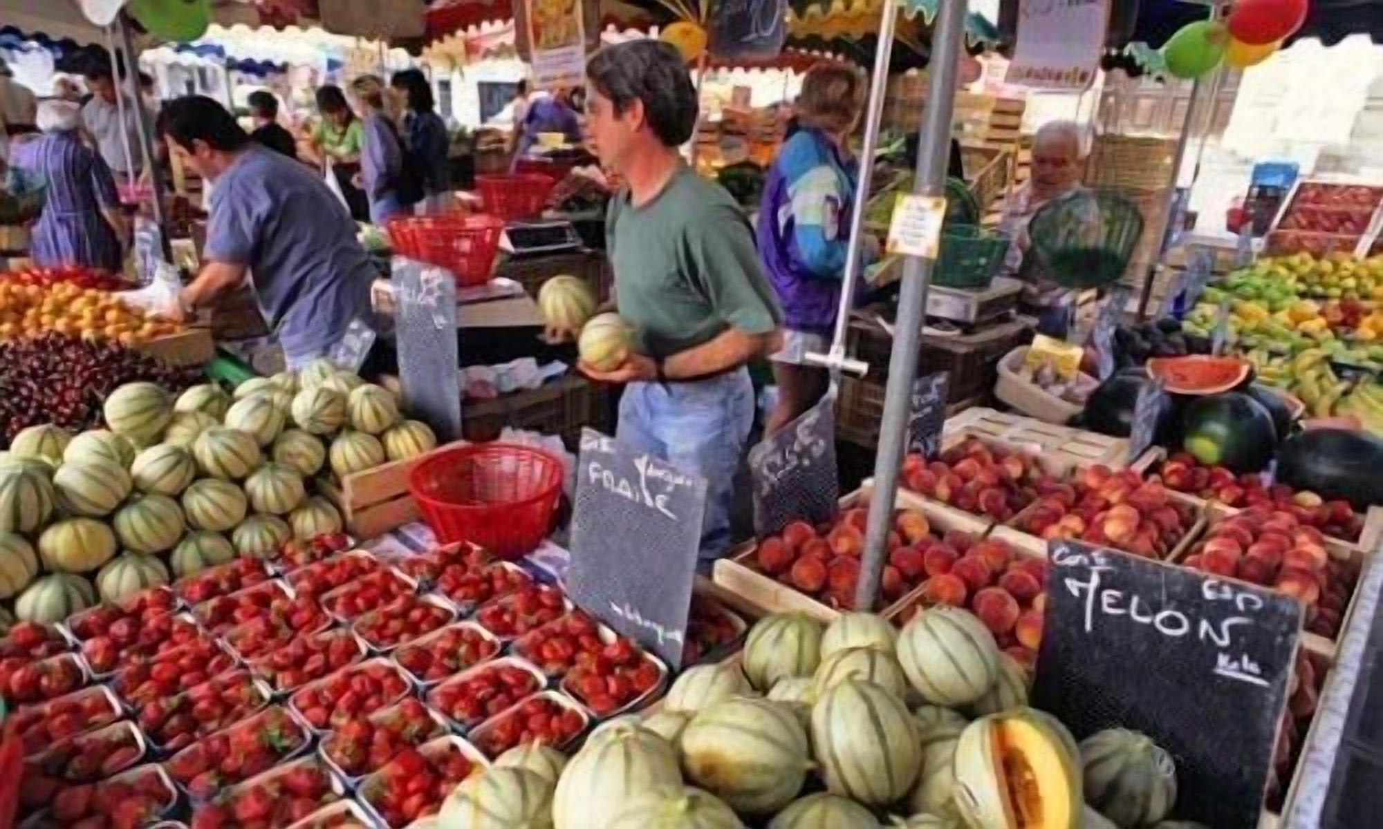 Un puesto de mercado con diversas frutas y verduras, incluyendo melones y fresas, y una persona sosteniendo un melón.
