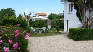 A garden with a gravel path leading to a white building with a door and windows. There are chairs and a table under a white umbrella.