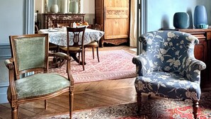 Ein Zimmer mit Esstisch, Stühlen, einem gemusterten Teppich, einer Holzkommode und einem blauen Sessel.