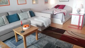 Un salon moderne avec un canapé gris, un petit lit agrémenté d’oreillers rouges, une table basse en bois avec des oranges, et une table d’appoint rouge et blanche.
