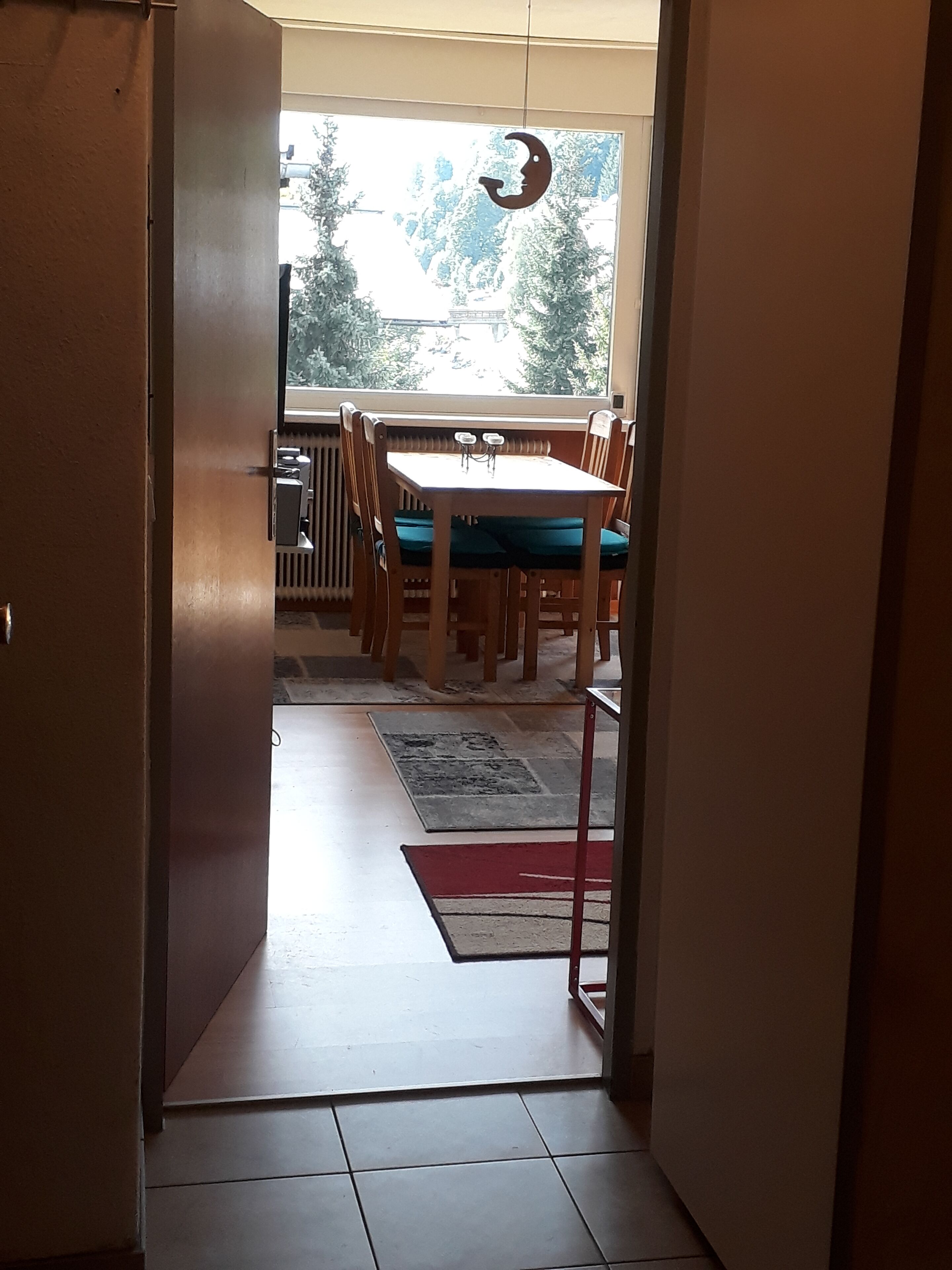 On aperçoit, à travers une porte, un coin repas avec une table et des chaises, un radiateur, et on peut voir des arbres à l’extérieur.