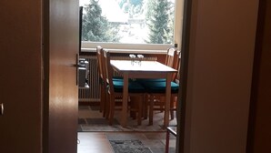 On aperçoit, à travers une porte, un coin repas avec une table et des chaises, un radiateur, et on peut voir des arbres à l’extérieur.